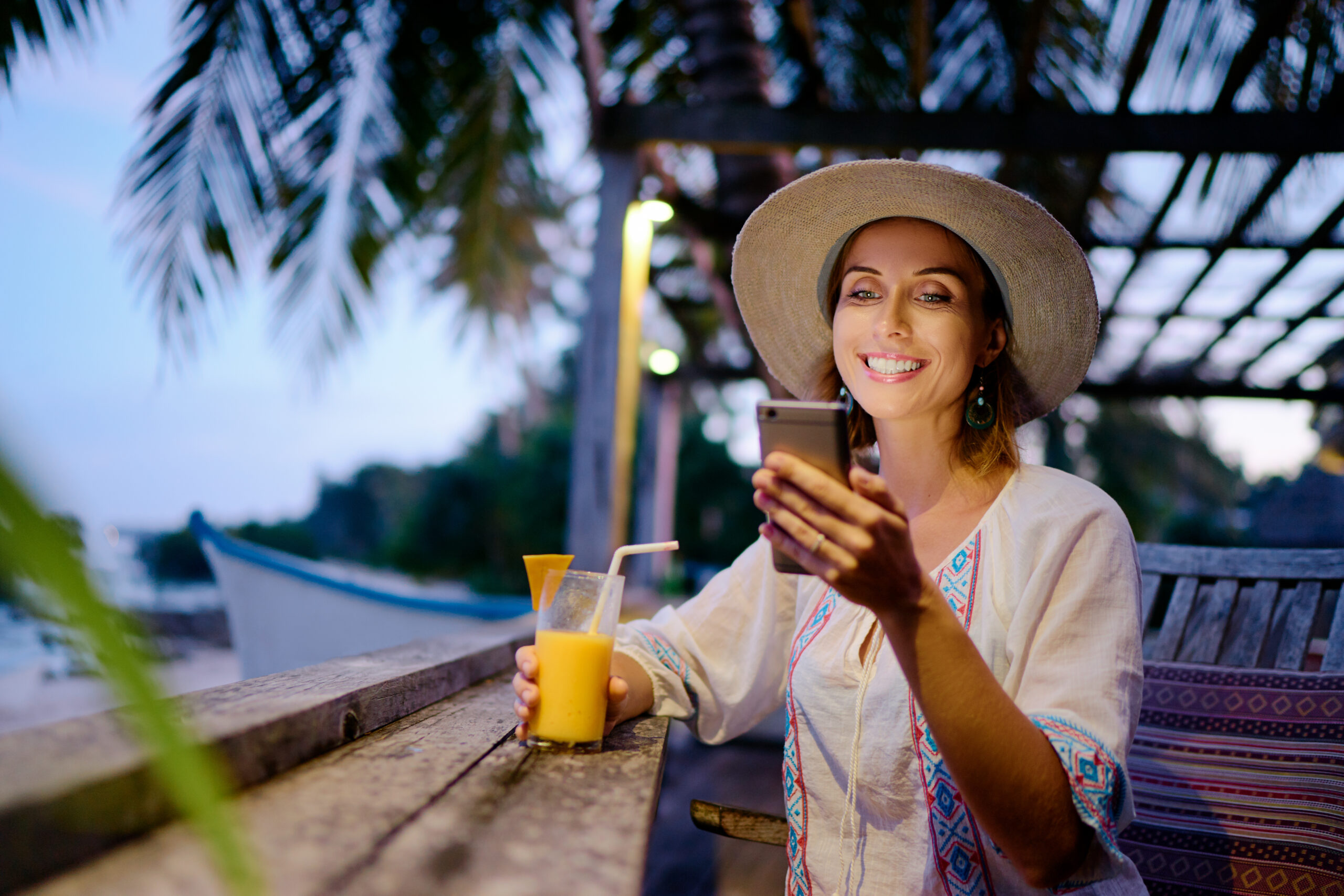 Young woman in a sun hat using her smartphone while sitting at a beachside cafe bar during a sunny day.
